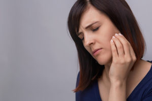 Young woman on grey background holding fingers to jaw in pain.